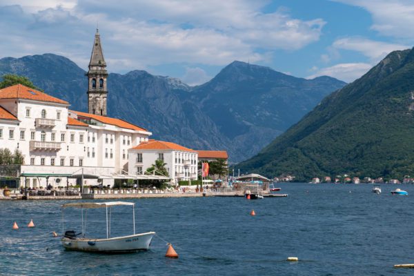 A scenic coastal town with historic architecture, a tall bell tower, surrounded by mountains, and boats on the calm blue water under a cloudy sky.