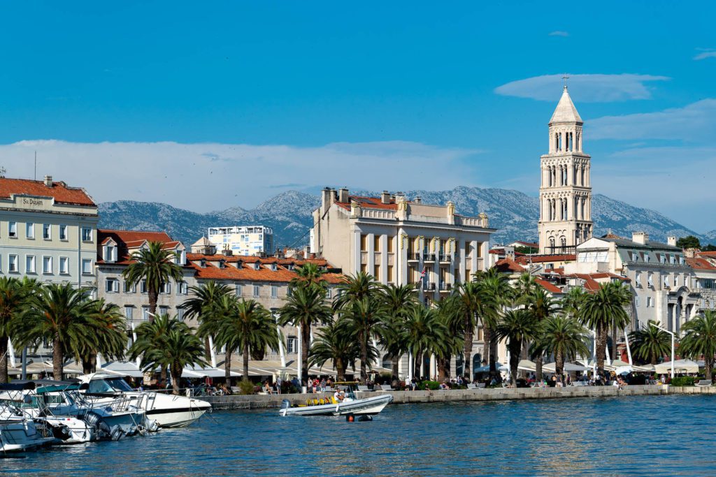 This image depicts a scenic waterfront lined with palm trees, boats, and historic buildings, featuring a prominent bell tower against a mountainous backdrop.