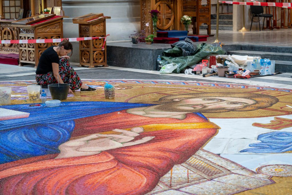 A person is creating a large, intricate floor mosaic of a religious figure inside a church, with colorful tiles and art supplies around them.