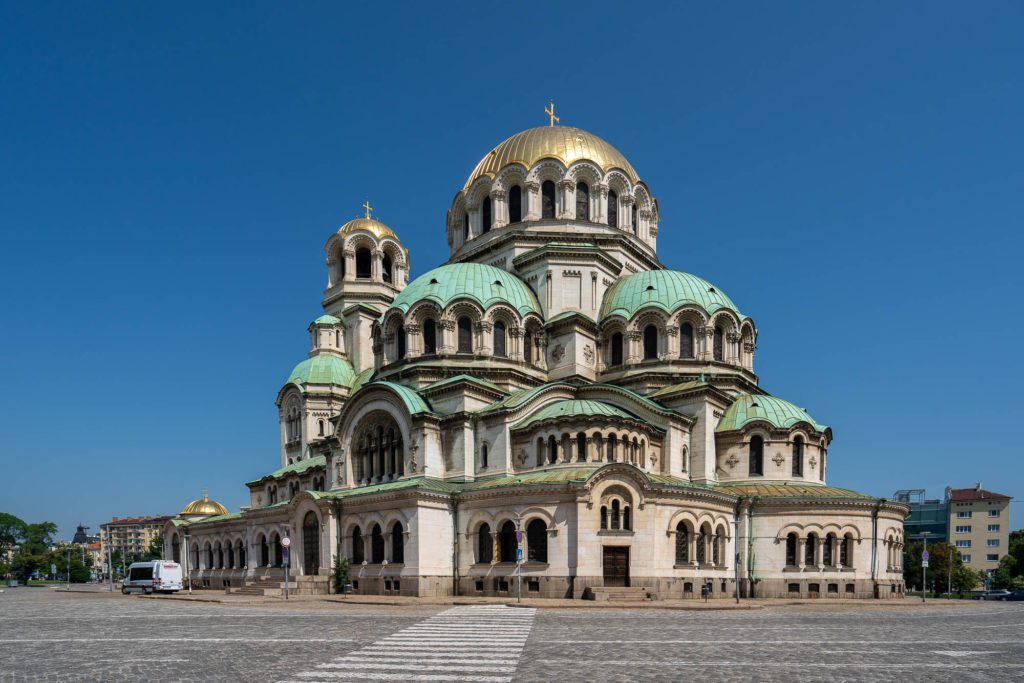 This image shows the Alexander Nevsky Cathedral in Sofia, Bulgaria. The large, ornate building features multiple domes and arches, with a clear blue sky above.