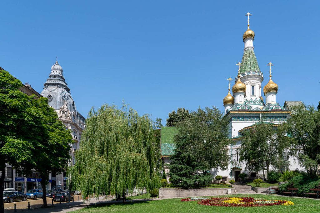 The image shows a vibrant urban park with lush trees, colorful flowerbeds, and a traditional Russian Orthodox church with onion domes and gold accents.