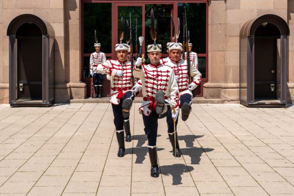 Three people in traditional ceremonial uniforms with stripes and medals march in formation, carrying rifles with bayonets. They are guarded by two individuals in similar attire.