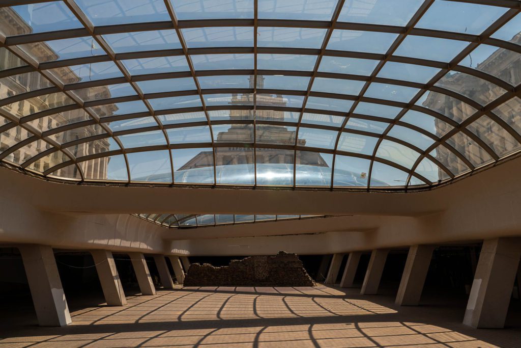 This image shows an interior space with a large glass dome ceiling, casting shadows on the floor. Sunlight filters through, highlighting geometric shapes and architectural details.