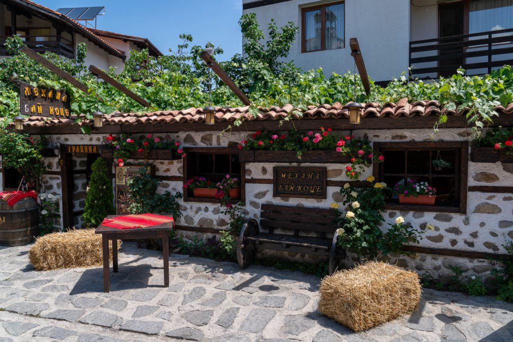 A quaint, rustic cafe with a cobblestone path, straw bales for seats, red cushions, climbing plants, and flowers adorning the traditional stone and wood facade.