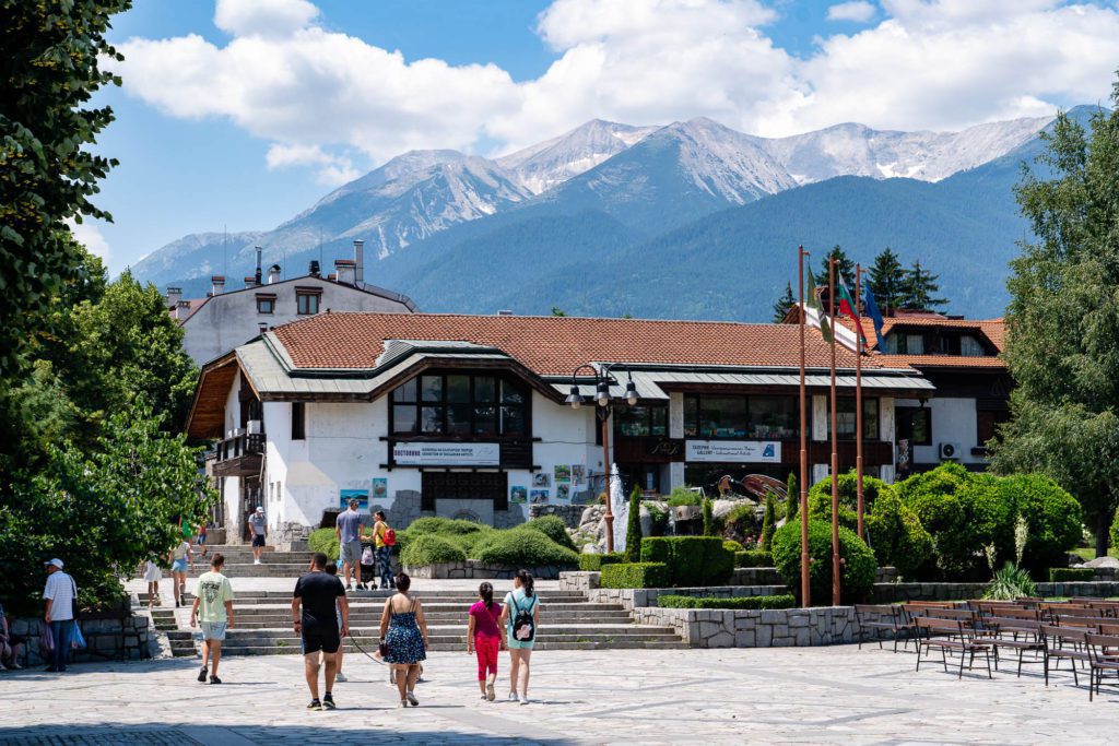 People walk in a sunny square with trees towards a traditional building with a backdrop of majestic mountains under a partly cloudy sky.
