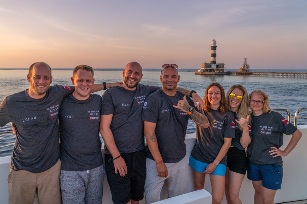 Seven people are posing on a boat at sunset, wearing matching t-shirts, smiling at the camera, with a lighthouse and the ocean in the background.