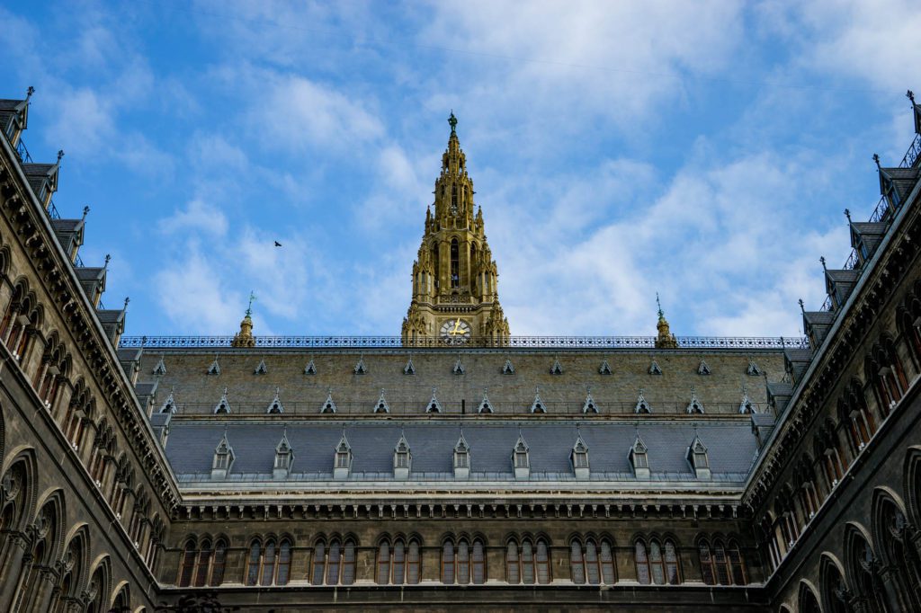 The image captures the ornate exterior of a Gothic-style building with a towering spire against a partially cloudy blue sky with a visible bird flying.