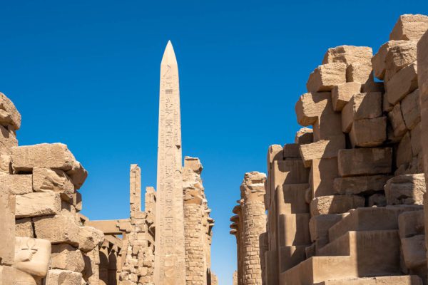 An ancient Egyptian temple complex with towering obelisk, hieroglyphs, and stone ruins under a clear blue sky, exhibiting historic architecture and archaeological significance.