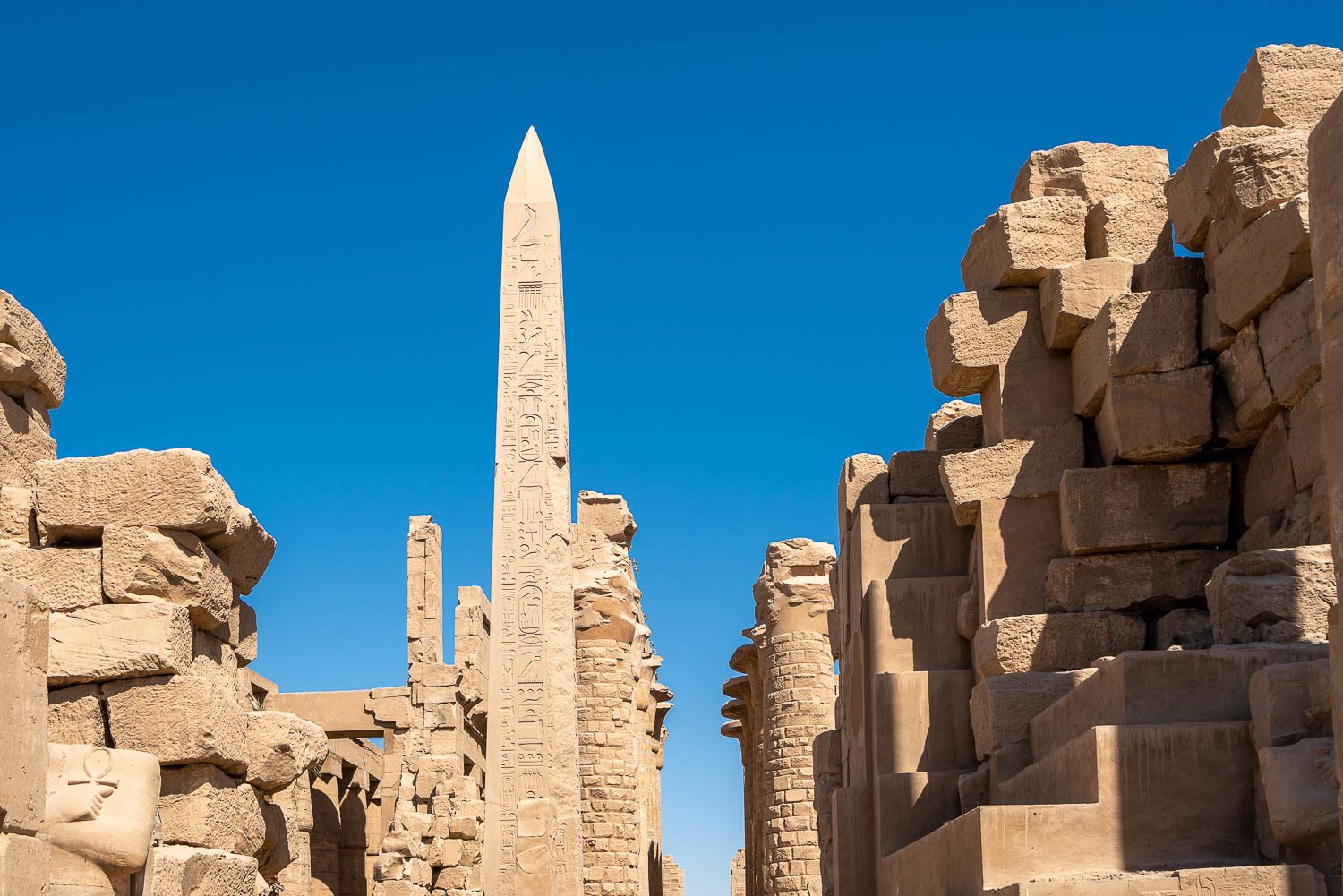 An ancient Egyptian temple complex with towering obelisk, hieroglyphs, and stone ruins under a clear blue sky, exhibiting historic architecture and archaeological significance.