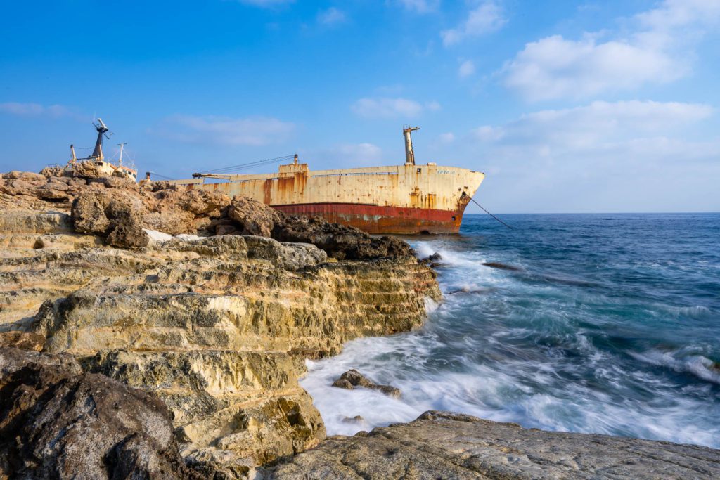 A large, rusted shipwreck is beached near rocky cliffs. Waves crash against the shore under a partly cloudy sky. A person stands at the cliff's edge.