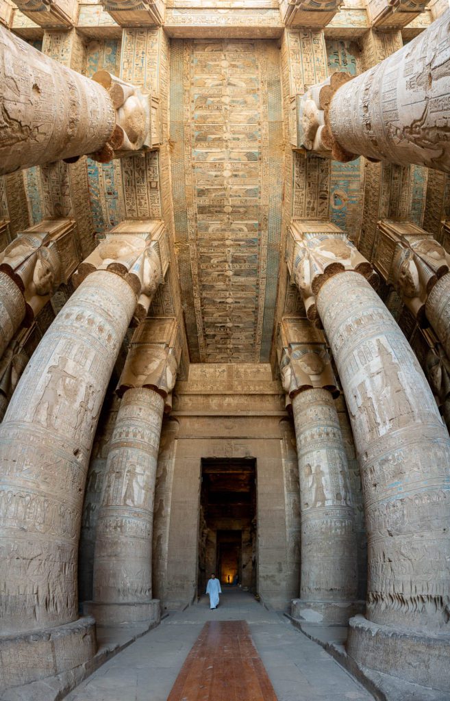 An ancient Egyptian temple interior with enormous columns, intricately carved with hieroglyphs and figures. A person in white walks down the central corridor.