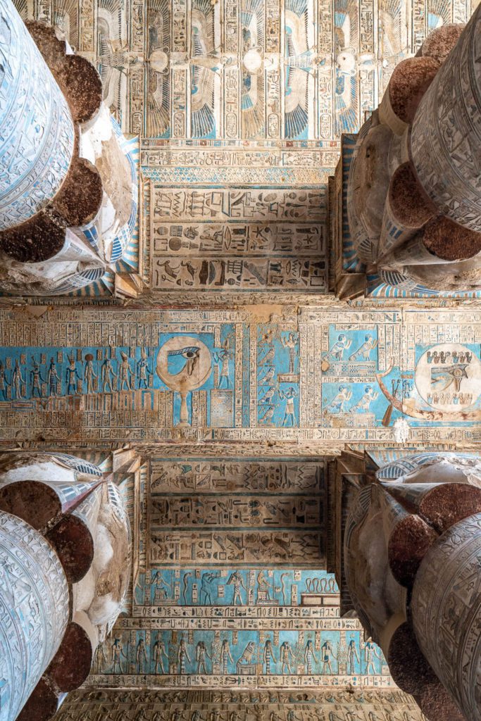 An ancient Egyptian temple ceiling adorned with intricate hieroglyphs and drawings, showcasing traditional motifs and deities, all in hues of blue, gold, and earthen tones.