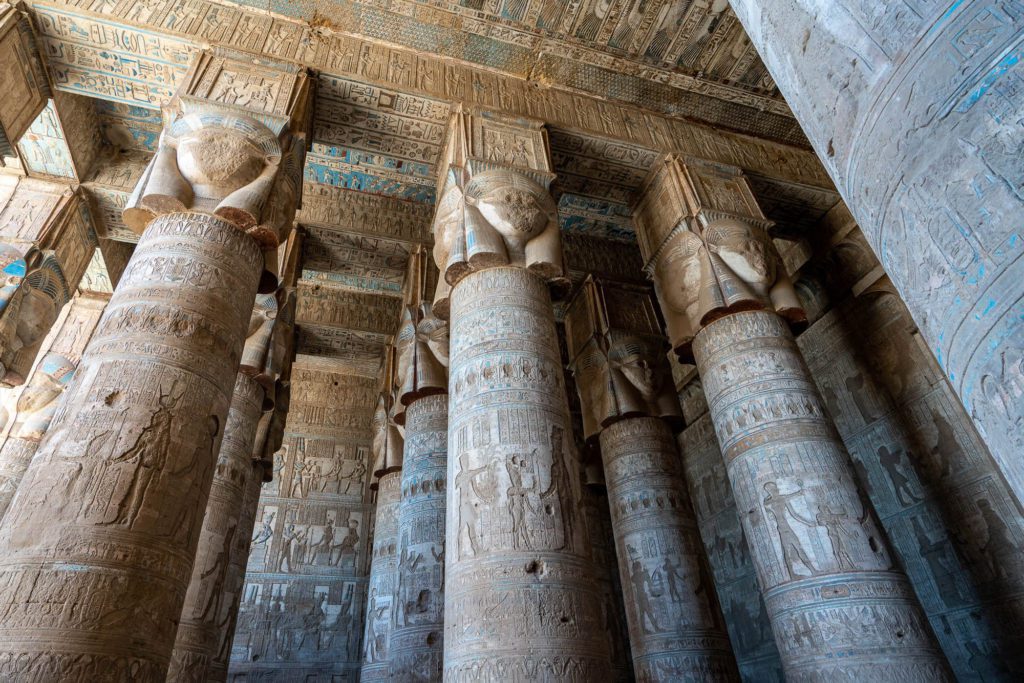 The image depicts ancient Egyptian temple columns adorned with hieroglyphics and intricate carvings, featuring a tall ceiling with remnants of colorful decorations.