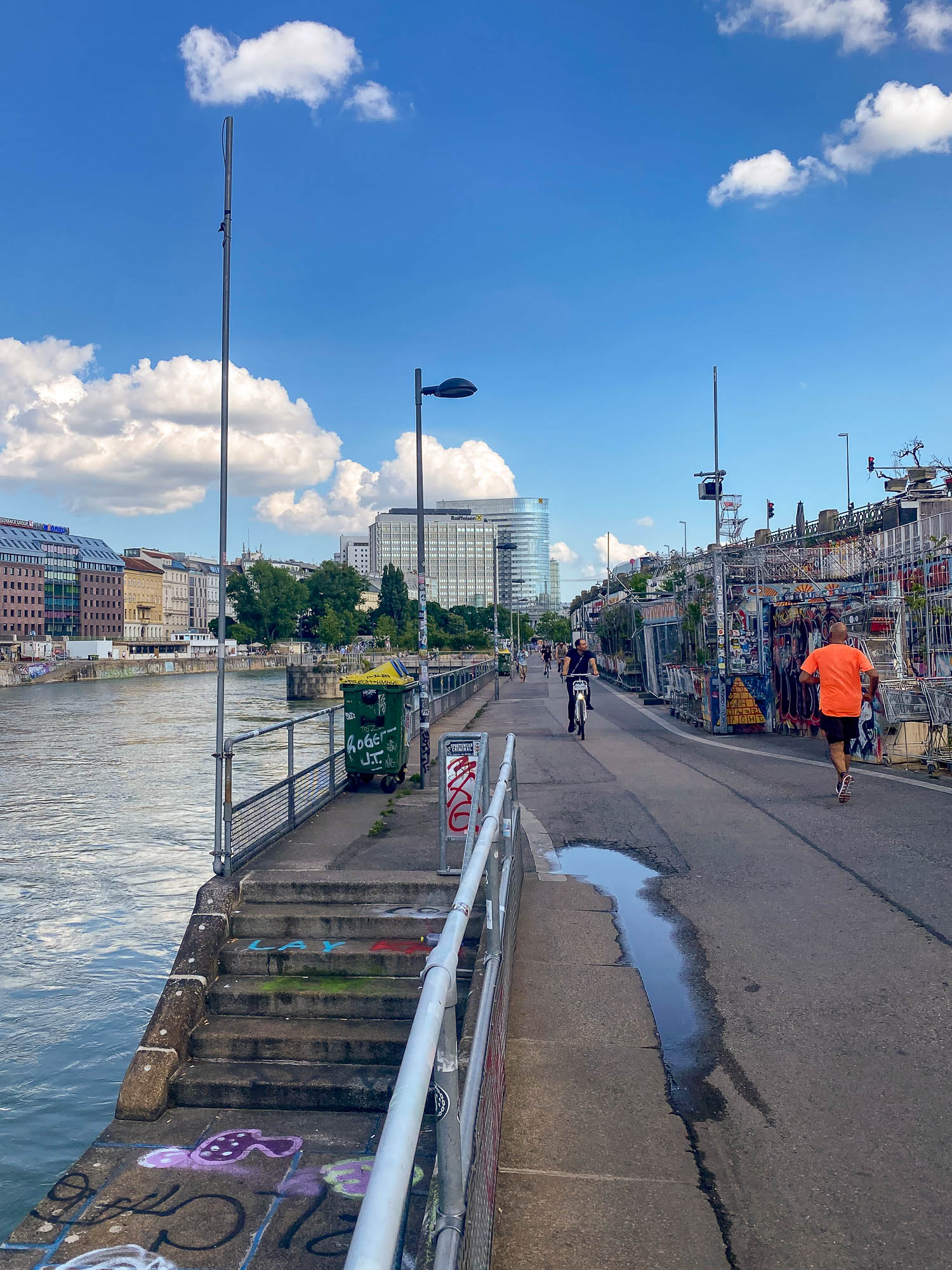 A riverside walkway with two people, one cycling and one walking. Graffiti, urban art, and market stalls line the path under a blue sky with clouds.