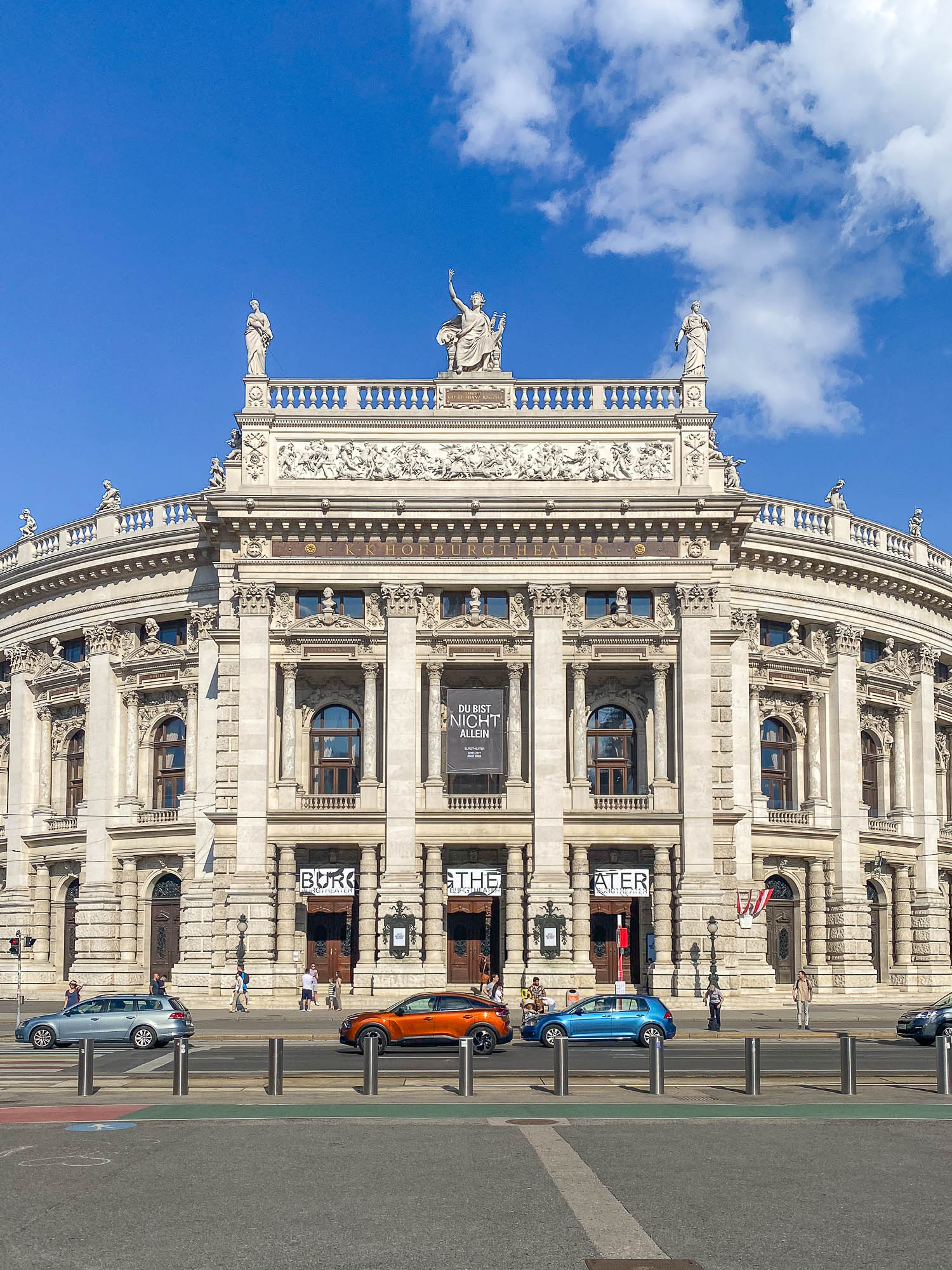 An ornate historical building with classical architecture under a blue sky with clouds. People and cars are visible in front, behind a metal barrier.