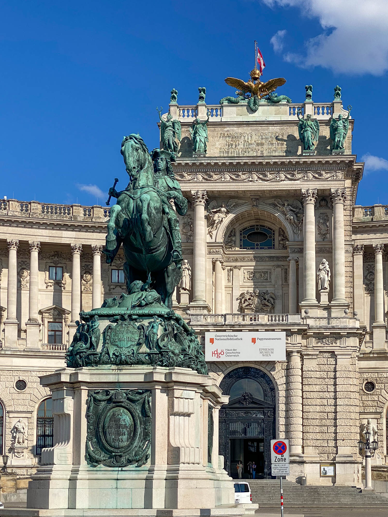 A bronze equestrian statue stands in front of an ornate building with sculptures, columns, and a winged figure atop its façade under a blue sky.