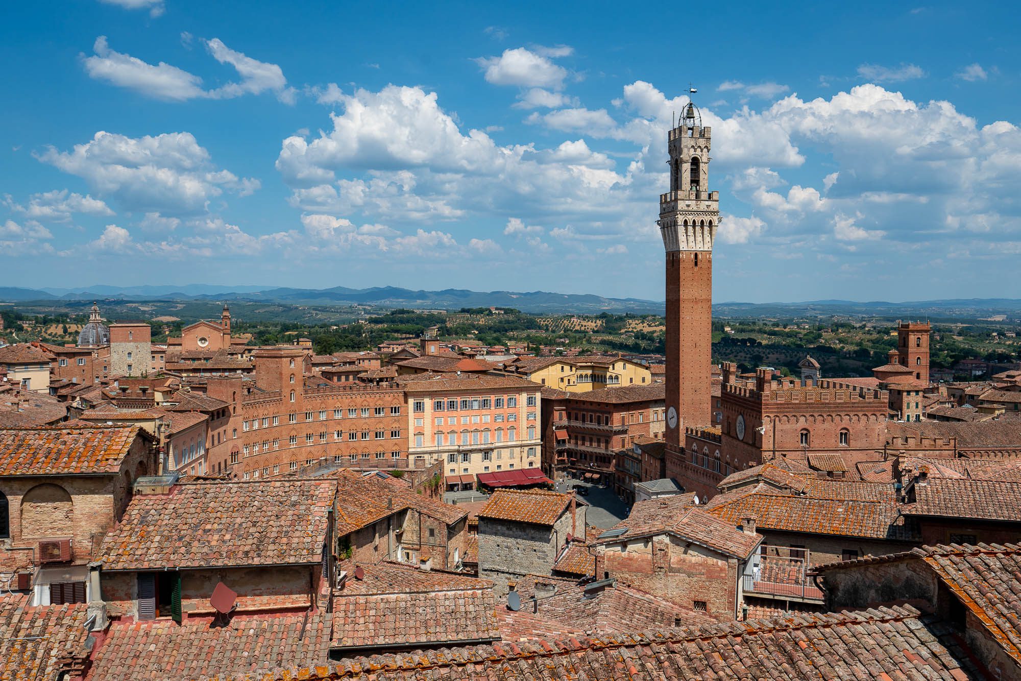 This is an aerial view of a historic Italian cityscape featuring terracotta rooftops, a prominent tower, and verdant rolling hills in the distance under a blue sky.