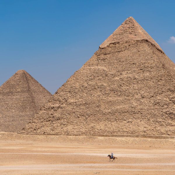 The image shows two ancient Egyptian pyramids under a clear blue sky, with a person riding a horse in the vast desert foreground.