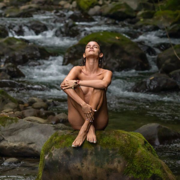 A person is sitting on a moss-covered rock, with eyes closed and knees drawn up, amid a serene stream with scattered rocks and flowing water.