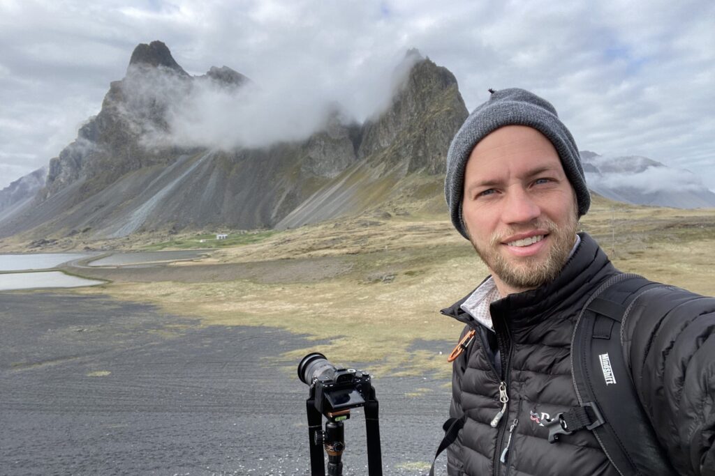 A person is taking a selfie with a scenic mountain background partly shrouded by clouds. A camera on a tripod stands in the foreground.