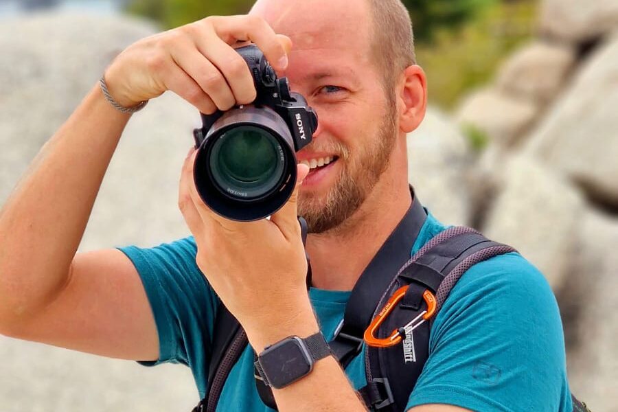 A smiling person with a bald head is taking a photograph with a DSLR camera outdoors while wearing a teal shirt and carrying a backpack.