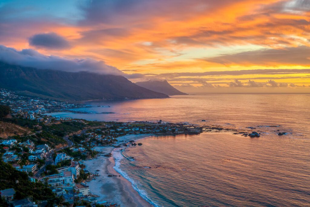 A coastal landscape at sunset with vibrant orange and blue skies, a mountain range shrouded in clouds, overlooking a serene beach and clustered buildings.