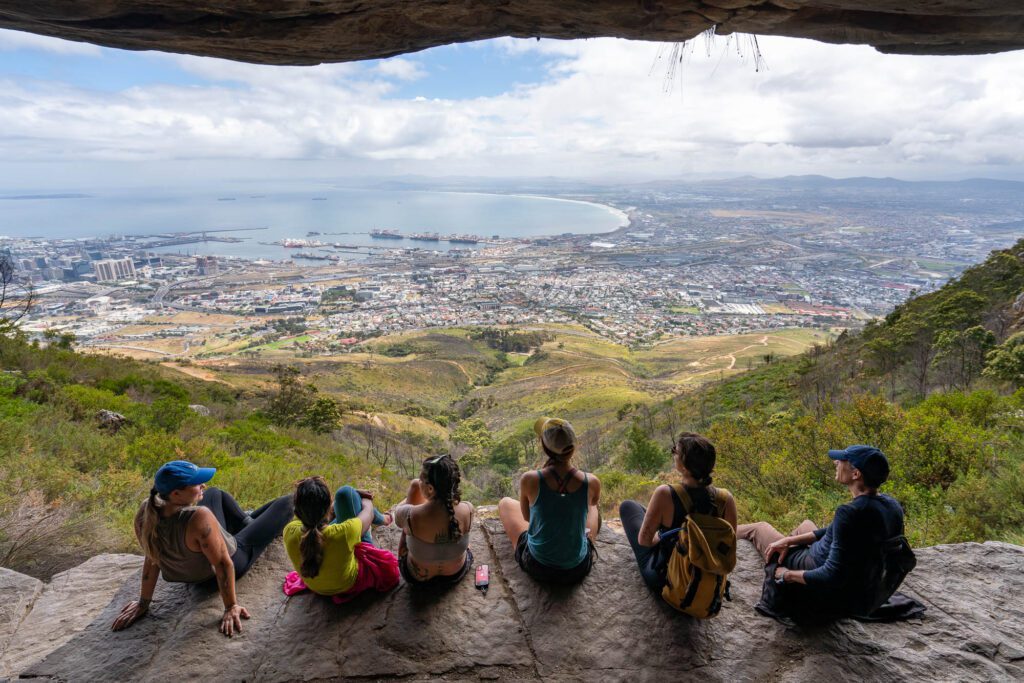 A group of people are seated on a rock ledge, overlooking a panoramic cityscape and coast from a high vantage point under an overhang.