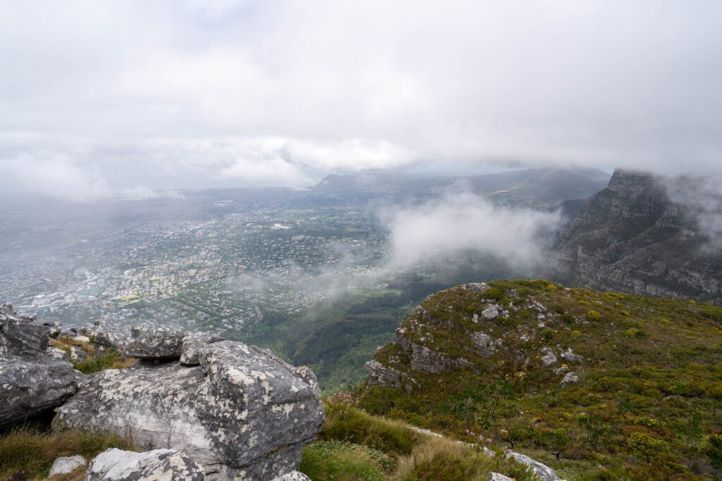 An expansive view from a mountain overlooking a valley, with low clouds or mist partially obscuring the landscape and residential areas below.