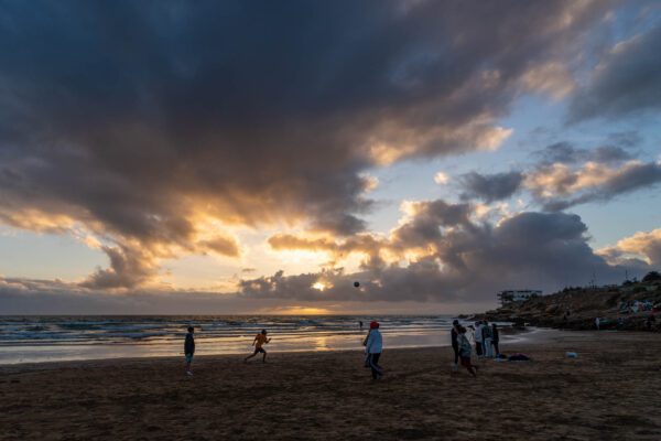 A beautiful beach scene at sunset with clouds in the sky. People play and walk on the sand. Warm colors reflect on the water.