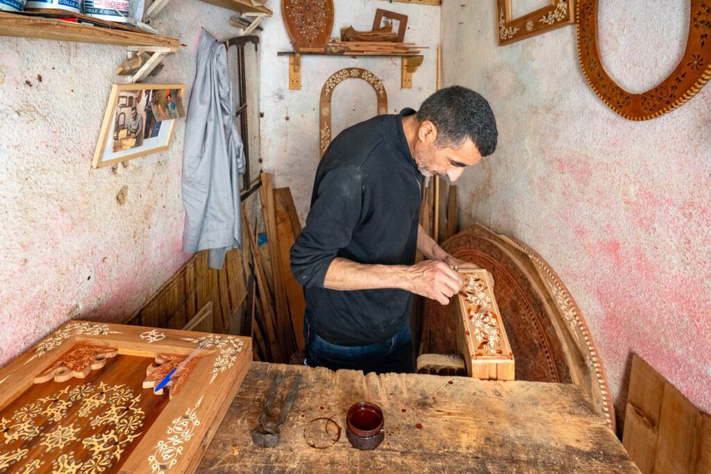 A person is meticulously working on decorative wooden objects in a workshop surrounded by tools and handmade items, indicating skilled craftsmanship in a traditional setting.