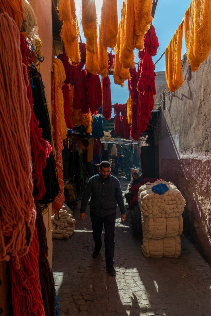 A person walks through a narrow alley lined with vibrant, freshly-dyed wool hanging to dry, creating a colorful canopy under a sunlit sky.