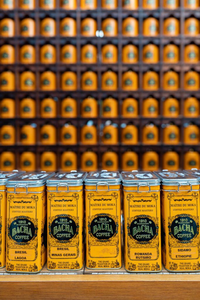 Rows of orange coffee canisters with black labels, displaying "BACHA COFFEE," are neatly arranged on wooden shelves in a gradient blur effect.