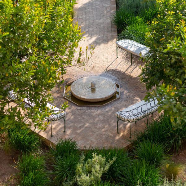 A peaceful garden with a circular path leading to a central fountain, surrounded by lush greenery and two white metal benches for rest and contemplation.