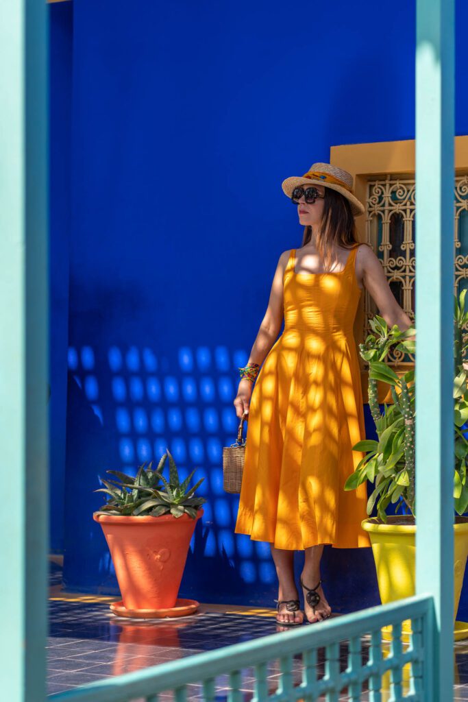 A person in a yellow dress and a straw hat stands amid vibrant blue walls, green plants, and terracotta pots, framed by a teal archway.