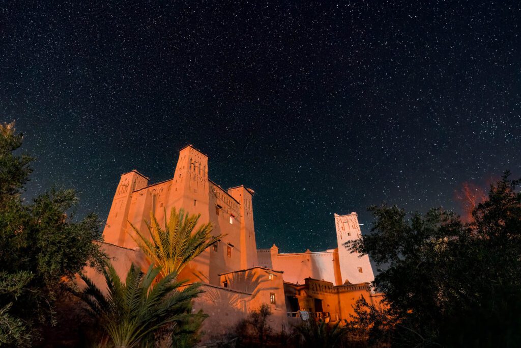 A star-filled night sky blankets an illuminated fortress-like building with tall towers. Silhouetted foliage adds tranquility to the serene, historical scene.