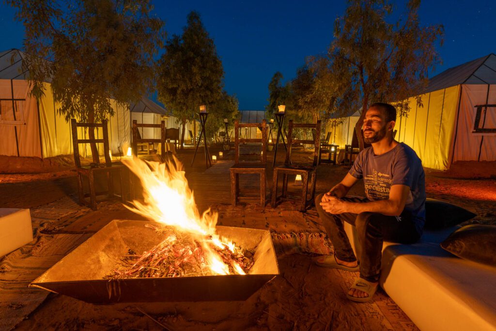 A person is smiling and sitting by a campfire at twilight, surrounded by tents and trees in what appears to be a glamping site.