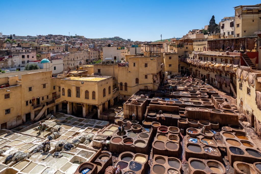 This image shows a traditional tannery with stone vats filled with dyes in a dense urban area. People are working amidst the vats under a clear sky.