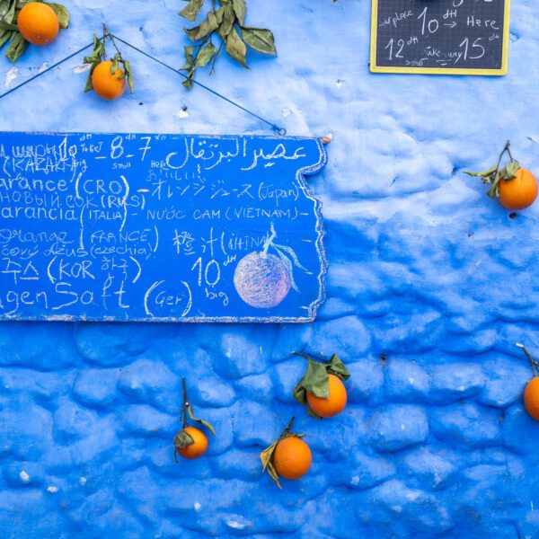 A blue textured wall displays oranges and price signs in various languages. The signs advertise fruit juice, showcasing multicultural communication and commerce.