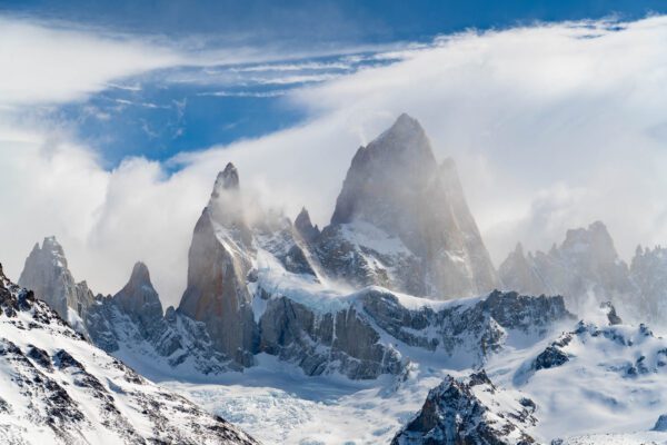 Majestic snow-covered mountain peaks rise sharply against a blue sky, with wispy clouds partially enveloping the rugged, icy landscape below.