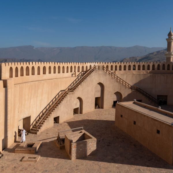 The image shows an ancient fort with crenellated walls, a minaret, and stairs leading upwards. It's a sunny day with clear blue skies and mountainous background.