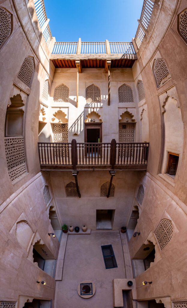 An architectural photo showcasing a traditional courtyard with balconies, ornate latticework, and wooden features under a clear blue sky. The design suggests Middle Eastern influences.