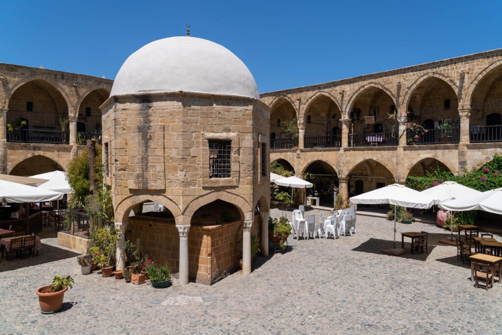 The image shows a historical stone building with arched corridors, a central dome, outdoor seating with umbrellas, and potted plants under a clear blue sky.