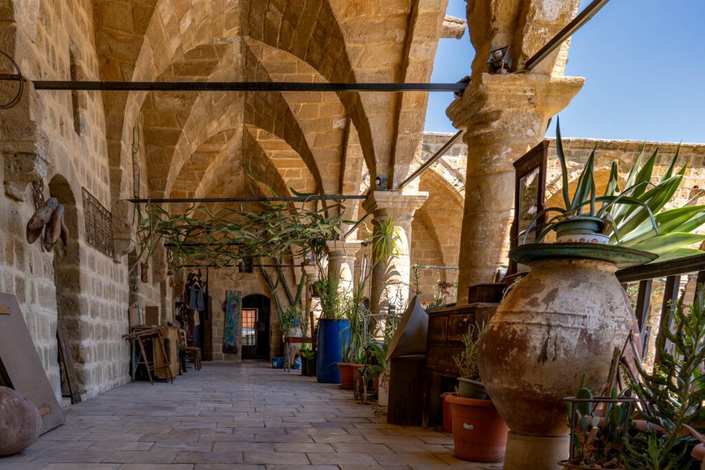 An old stone arcade with arched ceilings lined with potted plants, large clay jars, and rustic decorative items. Sunlight illuminates the tranquil corridor.