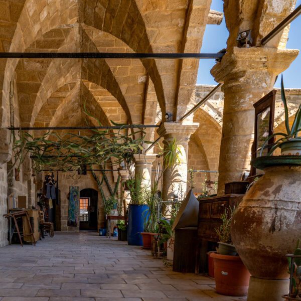 An old stone arcade with arched ceilings lined with potted plants, large clay jars, and rustic decorative items. Sunlight illuminates the tranquil corridor.