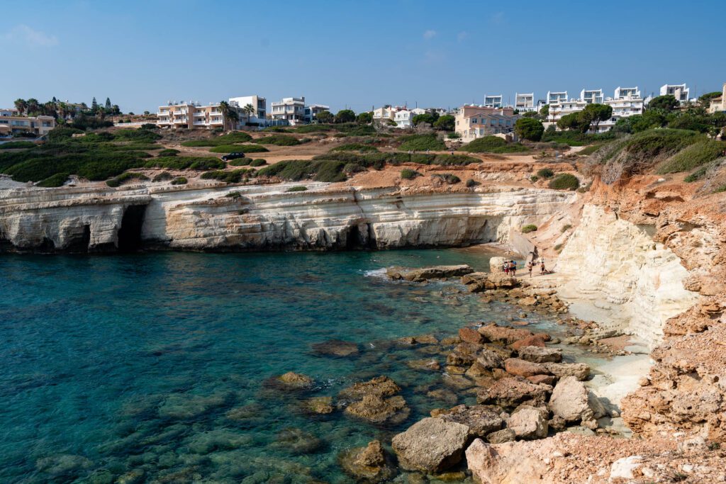 A scenic coastal landscape with clear turquoise waters, rocky cliffs, and buildings atop. People are by the water's edge enjoying the serene environment.