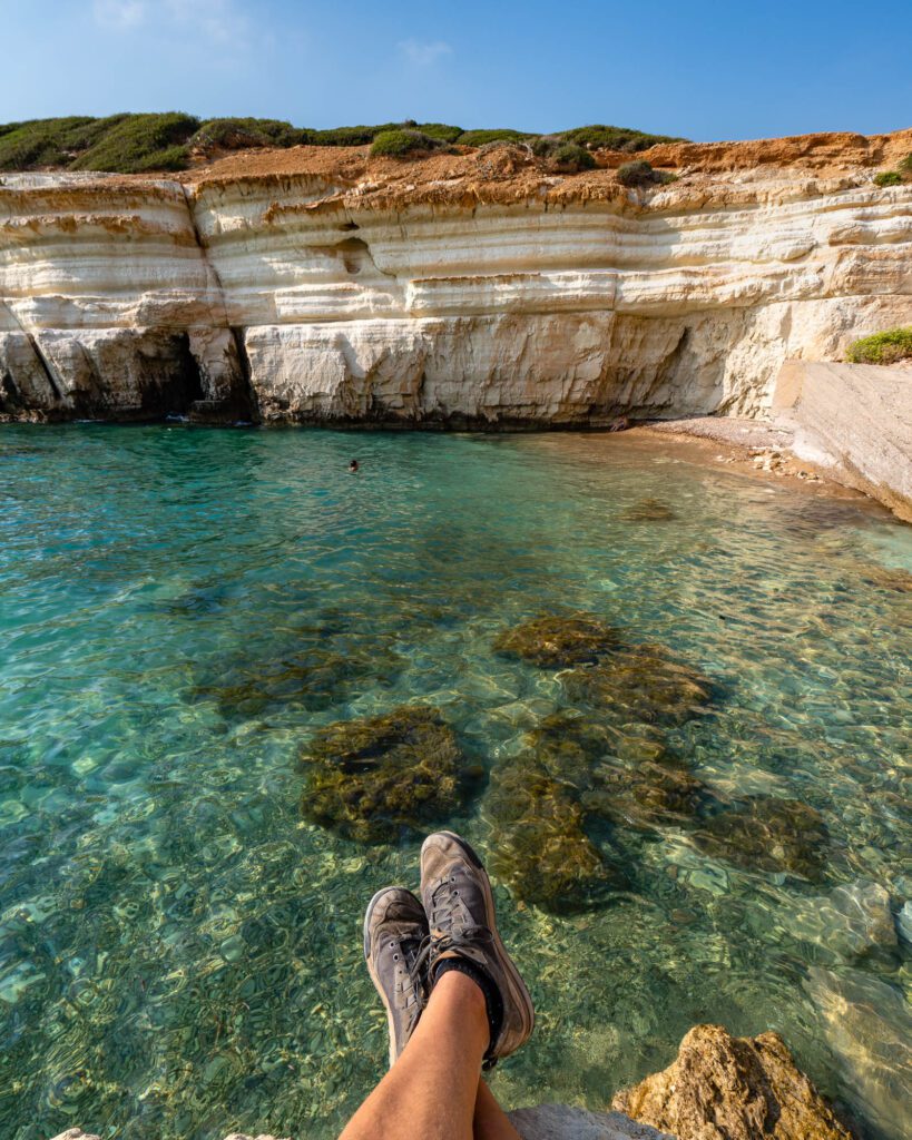 A person's legs are extended towards clear turquoise water in front of a layered rocky cliff under a blue sky, suggesting a relaxed viewpoint.