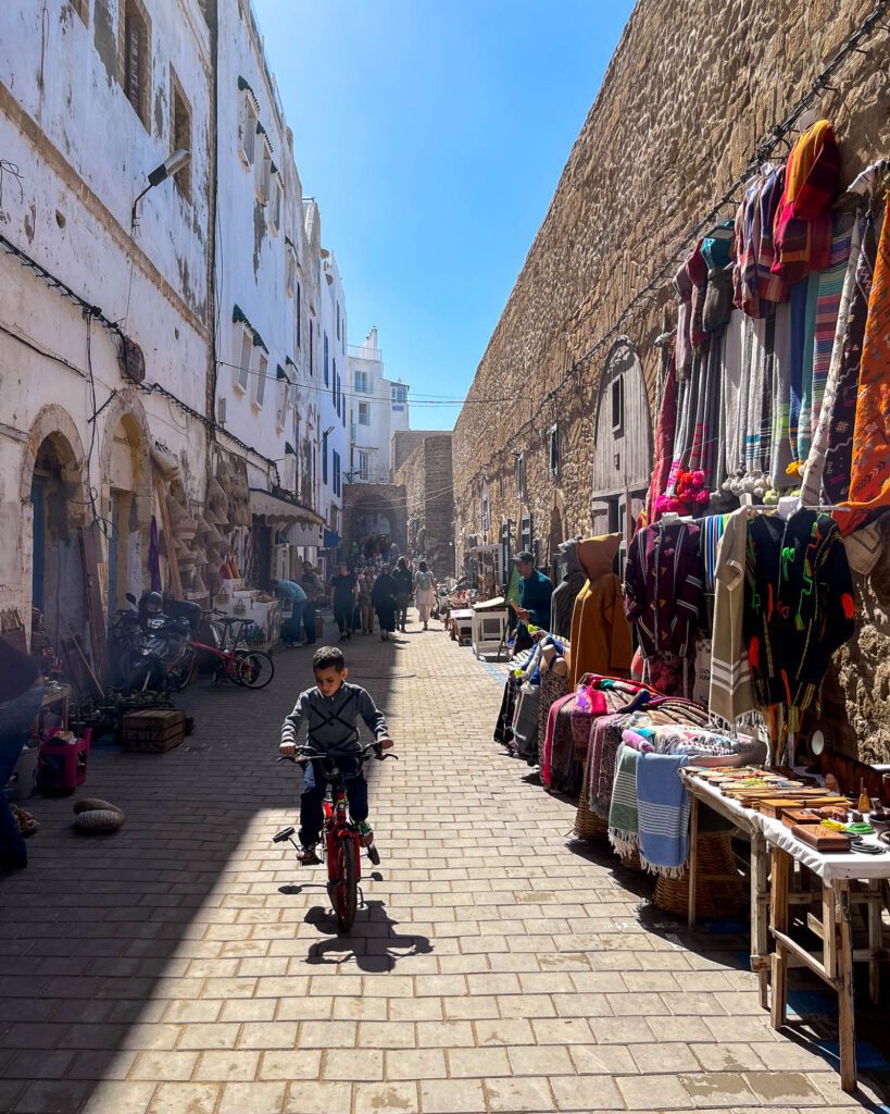A bustling market street lined with colorful textiles and wares. People browse items under the bright sunlight. A child on a bicycle adds to the scene's liveliness.