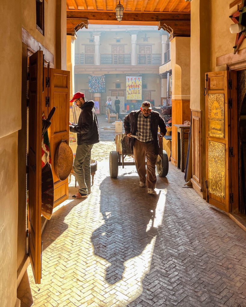 A person is pulling a cart in a sunlit traditional market alley with cobblestones, another person observes, surrounded by ornate doors and artisan goods.