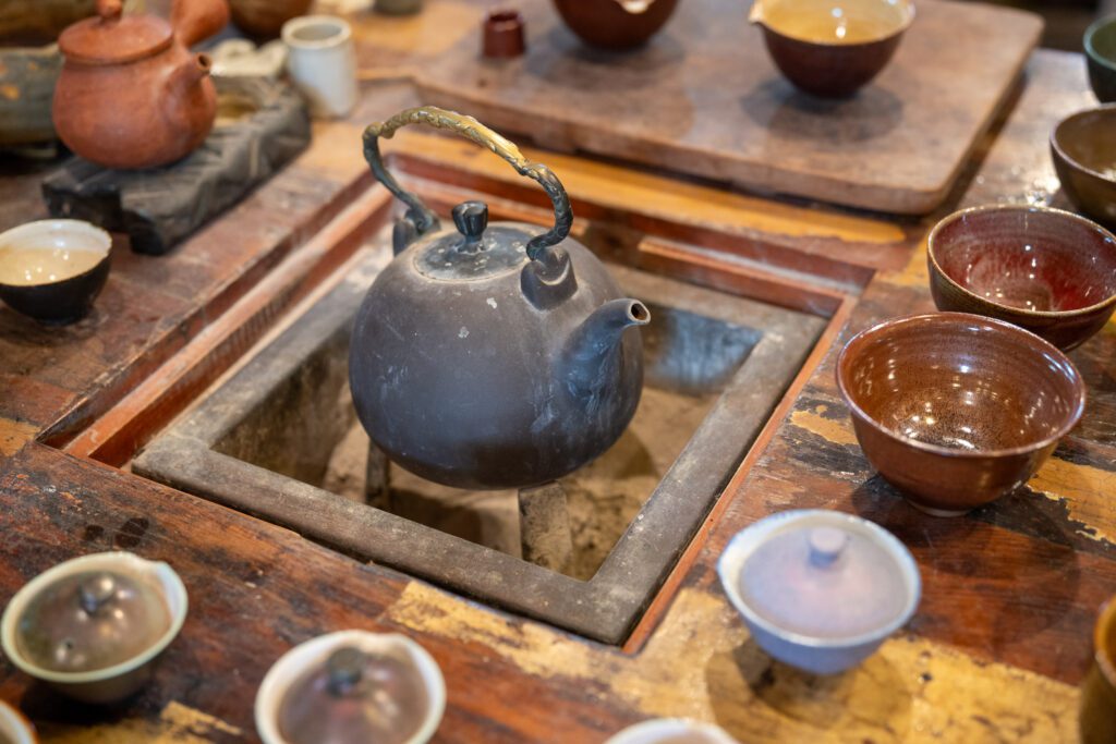 A rustic teapot sits on a stove surrounded by assorted ceramic cups in warm, earthy tones, creating a cozy, traditional tea setting.