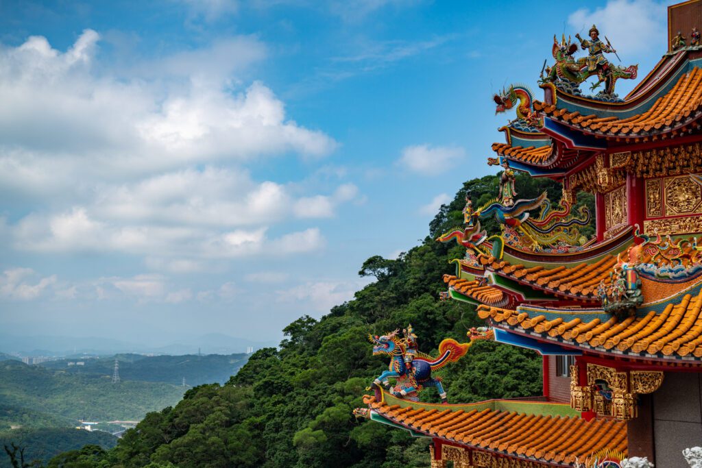 Traditional Chinese temple with ornate rooftop decorations, overlooking lush green mountains and a vast sky. No persons visible.